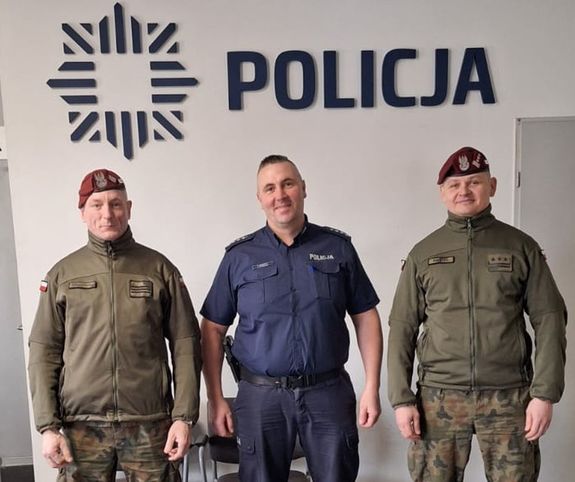 Policjant i funkcjonariusze wojska stoją ustawieni do zdjęcia, nad nimi jest napis Policja.
