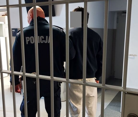 Policjant stoi obok zatrzymanego mężczyzny za okratowanymi drzwiami.