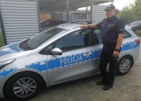 Umundurowany policjant w czapce stoi na tle policyjnego radiowozu