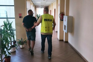 policjant w kamizelce z napisem Policja prowadzi zatrzymanego mężczyznę, który ma na rękach trzymanych z tyłu założone kajdanki