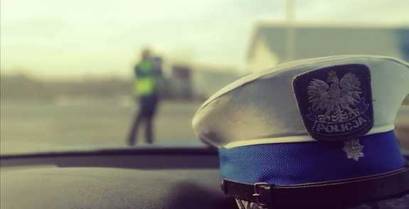 czapka policjanta ruchu drogowego w rozmytym tle widoczny policjant w odblaskowej kamizelce dokonujący pomiaru prędkości