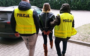 policjanci w kamizelkach odblaskowych prowadza do samochodu zatrzymaną kobietę, która na rękach trzymanych z tyłu ma nałożone kajdanki