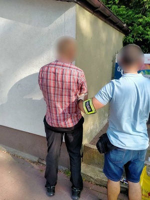Nieumundurowany policjant z opaską na lewym przedramieniu z napisem Policja, trzyma za prawe przedramię zatrzymanego mężczyznę, stojącego po jego lewej stronie. Obaj odwróceni plecami do obiektywu.