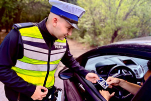 Umundurowany policjant podaje kierowcy naklejkę kampanii &quot;Mistrzu Zwolnij&quot;.