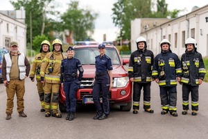 Umundurowane Policjantki i strażacy na tle pojazdu służbowego straży pożarnej.