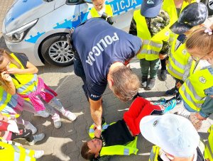 Pochylony policjant pokazuje dzieciom pierwszą pomoc przedmedyczną.