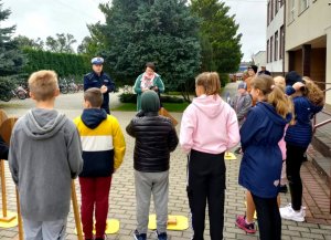 Umundurowany policjant ruchu drogowego, nauczycielka oraz młodzież podchodząca do egzaminu na kartę rowerową.