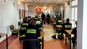 Policjant i policjantka, pies służbowy i strażacy ochotnicy siedzący w ławkach.