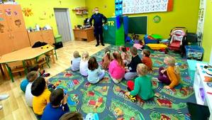 Policjant w mundurze stojący przed dziećmi w klasie, dzieci siedzą na dywanie.