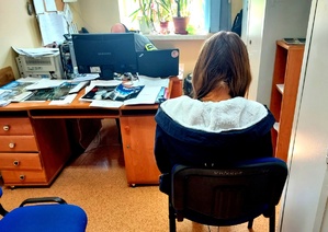 Kobieta siedząca tyłem na krzesełku, przed nią biurko z komputerem i dokumentami.