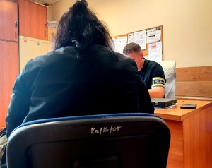 Kobieta siedząca na krzesełku przed nią przy biurku siedzi policjant  z opaską na ręce z napisem policja.