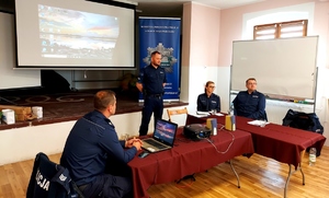 Policjanci w mundurach siedzący na krzesłach i jeden policjant stojący i rozmawiający z seniorami.