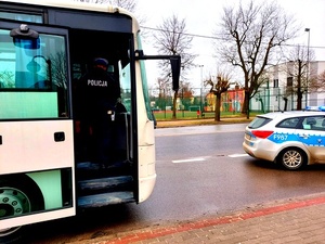 Stojący autokar, przed nim oznakowany radiowóz. Do autokaru wsiada umundurowany policjant.