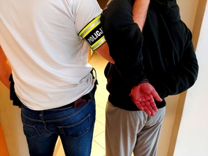 policjant w ubraniu cywilnym z opaską z napisem policja na ręce trzyma mężczyznę który ma kajdanki na ręce założone z tyłu obaj stoją tyłem.