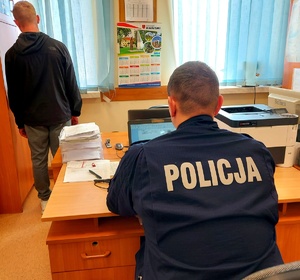 Policjant siedzący przy komputerze i mężczyzna stojący tyłem.