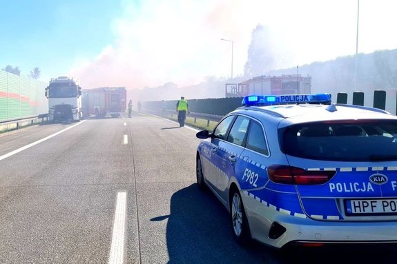 Radiowóz policyjny, policjant, ciężarówka, radiowóz straży pożarnej oraz dym z palącego się auta.