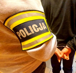Ręka z opaska z napisem Policja oraz postać stojąca tyłem z kajdankami na rękach.