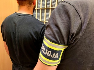 Mężczyzna stojący przy kratach i policjant w cywilnym umundurowaniu z opaska na ręce z napisem policja.