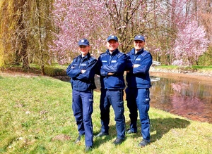 Troje policjantów, za nimi zbiornik wodny i kwitnące drzewa.