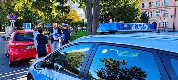 policjantka rozmawiająca z kursantami, instruktorem, radiowóz i samochód nauki jazdy.