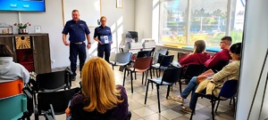 Policjant i policjantka z ulotkami stojący przed grupą osób.