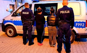 Policjanci stojący pob okach w środku obok siebie dwie kobiety na tle radiowozu policyjnego.