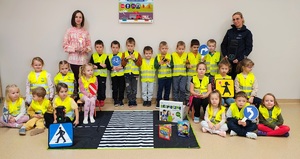 Policjantka, dzieci w kamizelkach odblaskowych trzymające znaki drogowe i nauczycielka na podłodze lezy mata z przejściem dla pieszych.