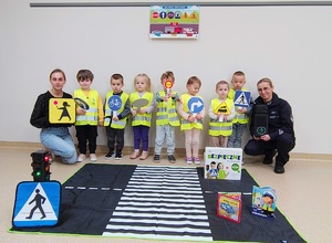 Policjantka, dzieci w kamizelkach odblaskowych trzymające znaki drogowe i nauczycielka na podłodze lezy mata z przejściem dla pieszych.