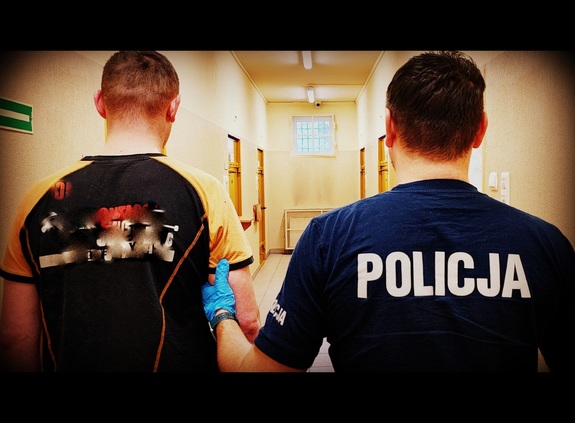 Policjant i zatrzymany mężczyzna stojący tyłem w korytarzu.