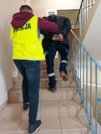 klatka schodowa komendy nieumundurowany policjant w żółtej kamizelce prowadzi zatrzymanego mężczyznę po schodach