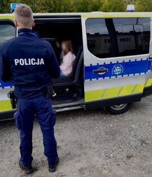 Dziewczynki siedzące w policyjnym radiowozie. Przy pojeździe stoi umundurowany policjant.