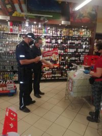 wizyta w sklepie, policjant dokonuj zapisuje w notatniku, członek komisji trzyma plakat, rozmawiają ze sprzedawcą