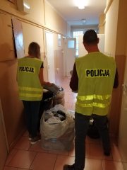 na korytarzu stojący tyłem policjant i policjantka ubrani z kamizelki odblaskowe z napisem policja wykonują oględziny odzieży , która znajduje się w workach,