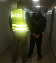 Na korytarzu w areszcie policjanta ubrany w kamizelkę odblaskowa z napisem POLICJA prowadzi zatrzymanego który ma założone kajdanki na ręce trzymane z tyłu, z prawej strony widać kratę na drzwiach