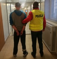 Na korytarzu w areszcie policjant ubrany w kamizelkę odblaskowa z napisem POLICJA prowadzi zatrzymanego który ma założone kajdanki na ręce trzymane z tyłu, z prawej strony widać kratę na drzwiach,