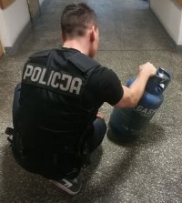 na korytarzu komendy policjant ubrany w czarną kamizelkę z napisem Policja  w kolorze białym wykonuje oględziny odzyskanej butli gazowej