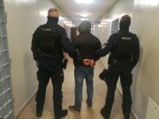 na korytarzu policyjnego aresztu umundurowani policjanci  prowadzą zatrzymanego, który ma założone kajdanki na ręce trzymane z tyłu, z boku po obu stronach  widać kraty