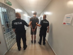 w areszcie dwóch umundurowanych policjantów prowadzą zatrzymanego który ma założone kajdanki na ręce trzymane z tyłu z lewej strony widać kratę na drzwiach