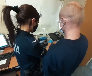 W pomieszczeniu służbowym umundurowana policjantka wykonuje odciski palców sprawcy rozboju. Policjantka która ma dłoniach rękawiczki  trzyma jego prawą dłoń, która przykłada do skanera. Mężczyzna stoi tyłem, ubrany jest w czarną bluzę, w pomieszczeniu widać sprzęt komputerowy stojący na biurku,