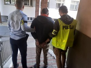 Po schodach prowadzących do aresztu dwóch policjantów z kotnych jeden ubrany jest w kamizelkę odblaskowa z napisem policja  prowadzą zatrzymanego który ma założone kajdanki na ręce trzymane z tyłu,