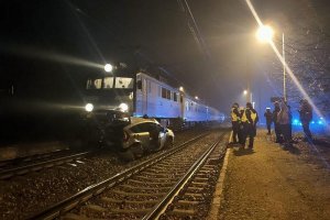 Na torach stoi pociąg który uderzył w samochód koloru srebrnego, auto jest uszkodzone  obok torów policjanci ubrani w kamizelki odblaskowe oraz stoi kilka osób