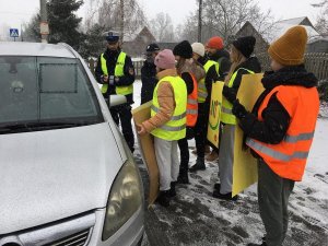 Dzieci wspólnie z policjantami przeprowadzają kontrole drogowe kierowców i wręczają woreczki  z ziemia  lub uśmiechnięte zawieszki, dzieci trzymaję w rękach plakat akcji