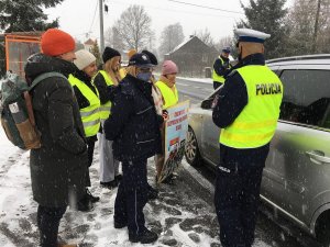 Dzieci wspólnie z policjantami przeprowadzają kontrole drogowe kierowców i wręczają woreczki  z ziemia  lub uśmiechnięte zawieszki, dzieci trzymaję w rękach plakat akcji