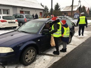 Dzieci wspólnie z policjantami przeprowadzają kontrole drogowe kierowców i wręczają woreczki  z ziemią  lub uśmiechnięte zawieszki, dzieci trzymają w rękach plakat akcji