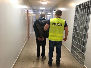 Na korytarzy aresztu nieumundurowany  policjant w kamizelce odblaskowej z napisem POLICJA prowadzi zatrzymanego