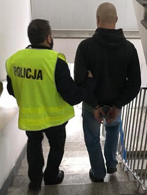 na schodach komendy policjant ubrany w kamizelkę odblaskową z napisem Policja prowadzi zatrzymanego który ma założone kajdanki na ręce trzymane z tyłu