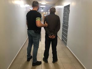 na korytarzu aresztu policjant ubrany w kamizelkę z napisem Wydział Kryminalny prowadzi zatrzymanego który ma złożone kajdanki na ręce trzymane z tyłu