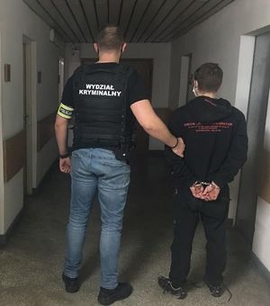 na korytarzu w budynku komendy  policjant ubrany w kamizelkę z napisem Wydział Kryminalny prowadzi zatrzymanego który ma złożone kajdanki na ręce trzymane z tyłu