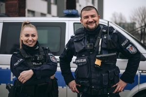 Przed radiowozem stoi umundurowana policjantka i policjant.