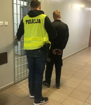 w areszcie policjant  prowadzi zatrzymanego, który ma kajdanki założone na ręce trzymane z tyłu